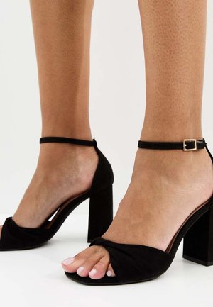 Dos pies con sandalias de tacón alto de ante negro, con correas en el tobillo y tacones cuadrados, sobre un fondo blanco.