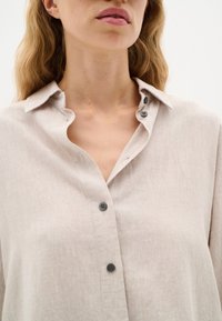 Light beige linen blouse with a folded collar, featuring dark buttons and a relaxed fit. The fabric has a subtle texture.