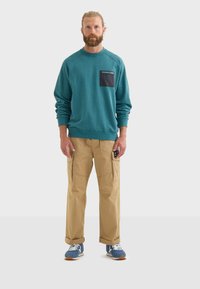 Sudadera verde azulado con un bolsillo negro y texto; pantalones cargo beige con bolsillos laterales; zapatillas azules con detalles blancos; estilo casual streetwear.