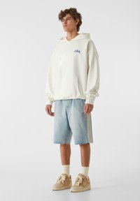 Balta kapuce ar priekšējo kabatu un zilu izšūtu logotipu, apvienota ar gaiši ziliem džinsu šortiem ar sānu svītrām un nolietotu apakšmalu.