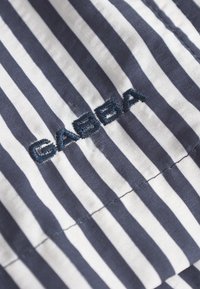 Tkanina s prugama u mornarsko plavoj i bijeloj boji, s izvezenim logotipom "GABBA". Vertikalne pruge s glatkom teksturom. Vidljivi detalji šavova.