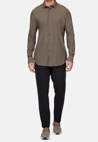 Camicia con bottoni in tessuto verde oliva scuro, con maniche lunghe, abbinata a pantaloni neri e sneakers beige, caratterizzata da un design attillato e da una trama liscia.