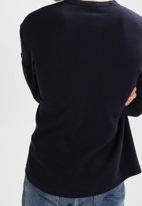 Person, der ein langärmliges, marineblaues Shirt und eine blauen Jeans trägt, von hinten gezeigt mit verschränkten Armen.