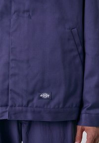 Marineblå arbeidsjakke laget av slitesterkt stoff, med et merke i nederkanten, sidelomme og rett kutt-design med minimale sømdetaljer.