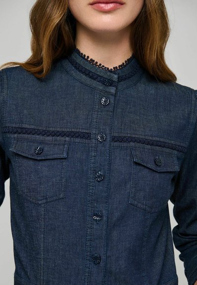 Femme portant une chemise en denim bleu foncé avec des boutons décoratifs et une bordure en dentelle sur le col et les poches poitrine, vue des épaules aux lèvres.