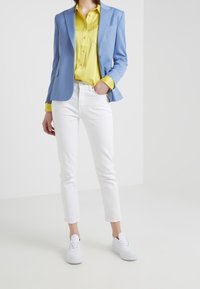 Blazer azul, camisa amarilla, jeans blancos y zapatillas blancas. El blazer tiene solapas con muesca; la camisa tiene un frente con botones y un bolsillo.