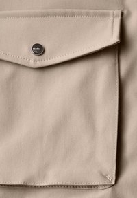 Tissu beige avec une surface texturée, avec une poche à rabat fermée par un bouton-pression métallique, des bords soigneusement cousus et un design minimaliste.