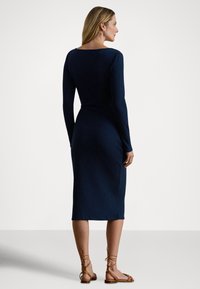 Marineblå ribbestrikket kjole med lange ermer og en tettsittende silhuett, med rund utringning bak og ankel-lang kant, kombinert med brune sandaler.