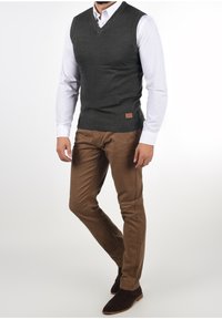 Veste en tricot gris charbon à col en V, portée sur une chemise blanche ; associée à un pantalon marron slim et des bottines foncées. Étiquette en cuir en accent sur la veste.