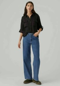 Schwarzes Hemd mit hochgekrempelten Ärmeln, kombiniert mit hochgeschnittenen blauen Jeans und grünen Plateau-Stiefeln, vor einem neutralen Hintergrund.