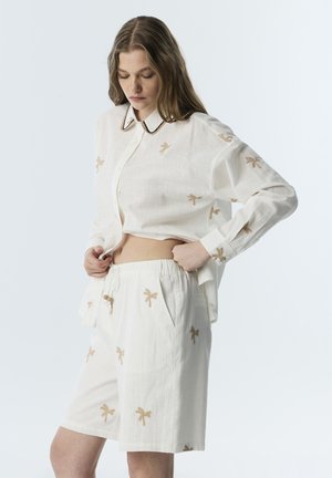 Vrouw draagt een losse witte blouse en bijpassende korte broek met beige geborduurde palmboompatronen, past de tailleband aan en kijkt naar beneden.