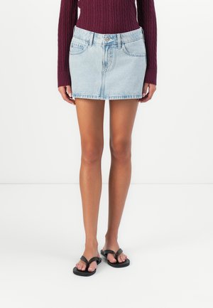 Kvinne iført en lyseblå denim miniskjørt, mørk lilla kabelstrikket genser og svarte flip-flop sandaler som står mot en hvit bakgrunn.