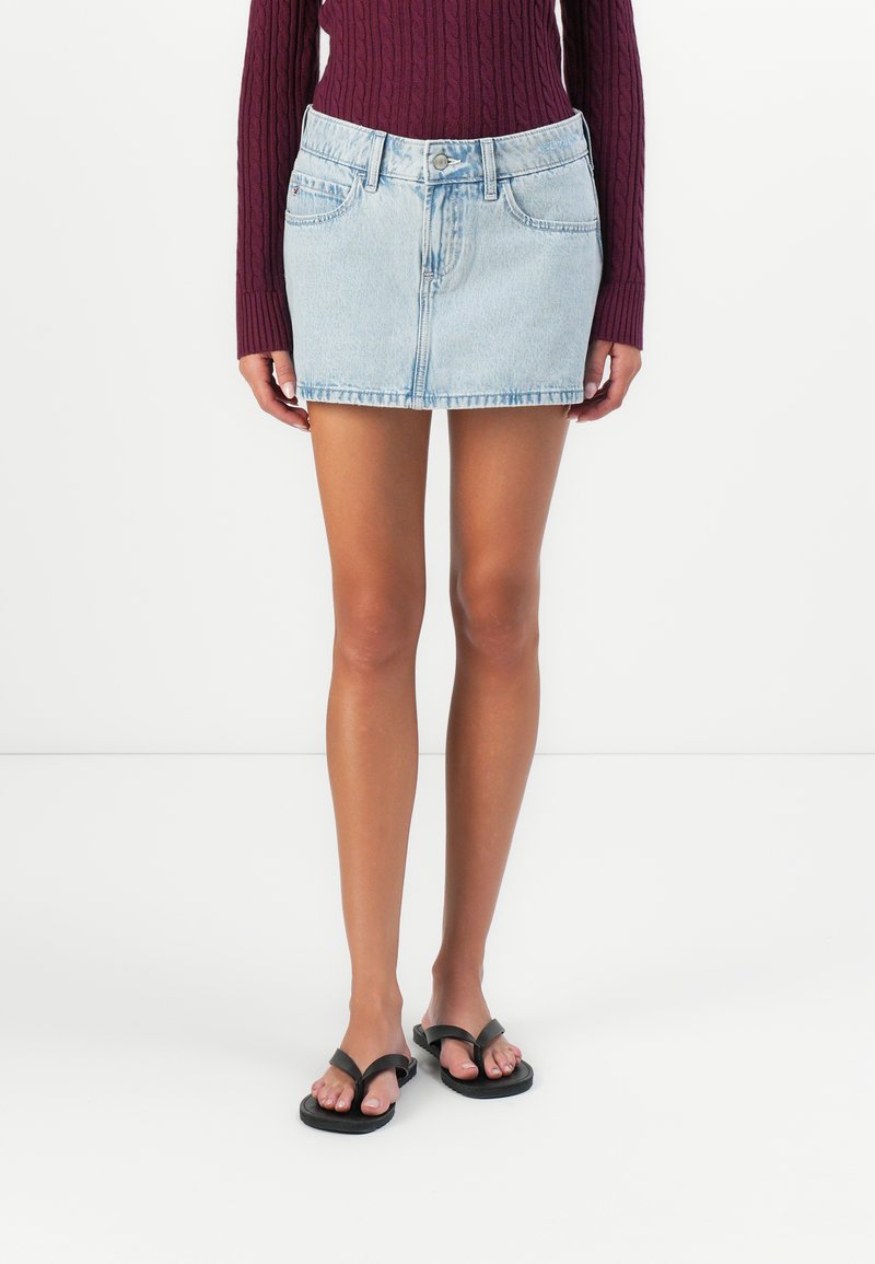 Kvinne iført en lyseblå denim miniskjørt, mørk lilla kabelstrikket genser og svarte flip-flop sandaler som står mot en hvit bakgrunn.
