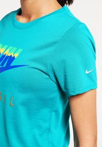 Camiseta Nike turquesa de manga corta, con un diseño gráfico colorido y letras doradas. El tejido parece suave y ligero.