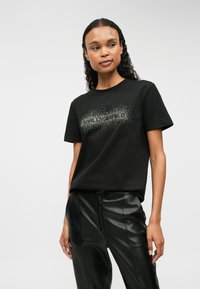 Melna kokvilnas T-krekls ar mirdzošu sudraba grafiku, kurā redzams teksts "CARL LAGERFELD", īsām piedurknēm un klasisku apaļu izgriezumu.