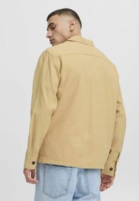 Chemise légère de couleur marron clair avec un col, des manches longues et des poignets à boutons. Le tissu a une texture lisse et un motif de tissage subtil.