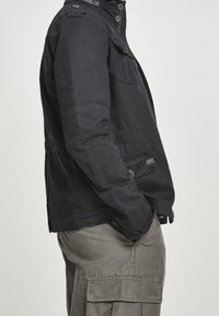 Svart, lättviktsjacka med strukturerad design, utrustad med hög krage, flera fickor och knapplister. Bärs över grå cargobyxor.