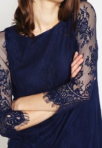 Mörkblå klänning med nätspetsärmar som har blommönster och fransdetaljer. Korsetten har en texturerad yta för extra djup.