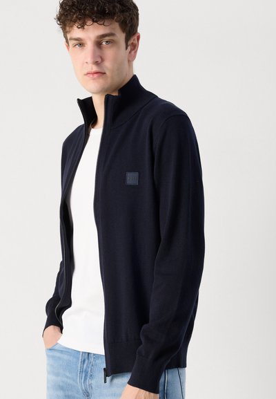 El abrigo de punto azul marino con cremallera presenta un cuello alto, puños acanalados, un pequeño parche con logo y una textura suave. Llevado sobre una camiseta blanca.