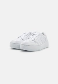 Baskets basses blanches avec un bout perforé, un design à lacets, une semelle rembourrée, une semelle extérieure en caoutchouc texturé et un branding minimaliste sur le côté.