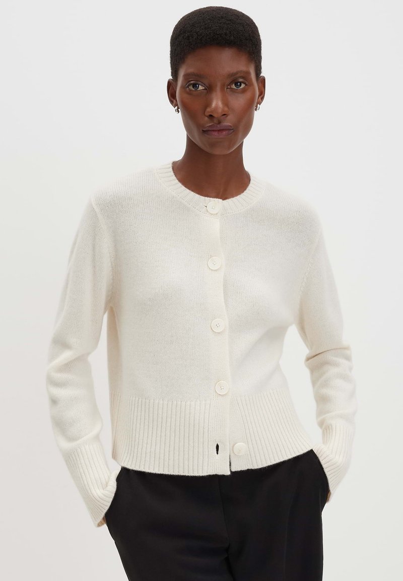 Ein Model trägt eine weiße, geknöpfte Strickjacke mit gerippten Bündchen und Saum, kombiniert mit schwarzen Hosen, und steht vor einem schlichten, hellen Hintergrund.