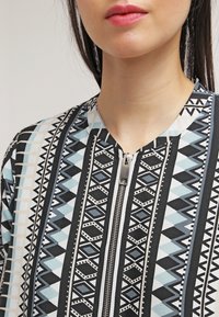 Ritssluiting aan de bovenkant met een geometrisch patroon in zwart, wit en blauw. Heeft een ronde halslijn en een gladde, gestructureerde stof.