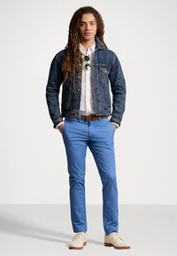 Μπουφάν από τζιν, λευκό πουκάμισο, μπλε παντελόνι και μπεζ παπούτσια· μακριά σγουρά μαλλιά, γυαλιά ηλίου, σε όρθια στάση μπροστά σε απλό φόντο.