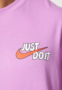 Krátký rukáv, světle fialové bavlněné tričko s textem "JUST DO IT" v bílé a oranžové barvě a logem zaškrtnutí. Hladká textura.