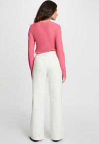 Femme de dos, portant un haut rose à manches longues et un pantalon large taille haute blanc, sur fond uni.