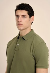 Camisa polo verde oliva hecha de un tejido texturizado, con cuello, dos botones y un pequeño logo en el pecho. Diseño ajustado.