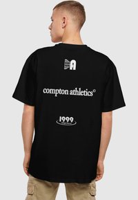 Fekete pamut póló laza szabással. Fehér felirat: "compton athletics", egy földgömb design, és az alján a "1999 Minden jog fenntartva" felirattal.