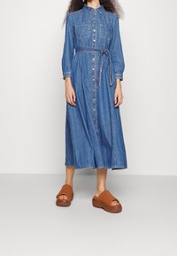 Denim shirt dress, long sleeves, button-down front, two chest pockets, waist tie, mid-calf length, finished with contrasting stitching; worn with brown platform sandals.