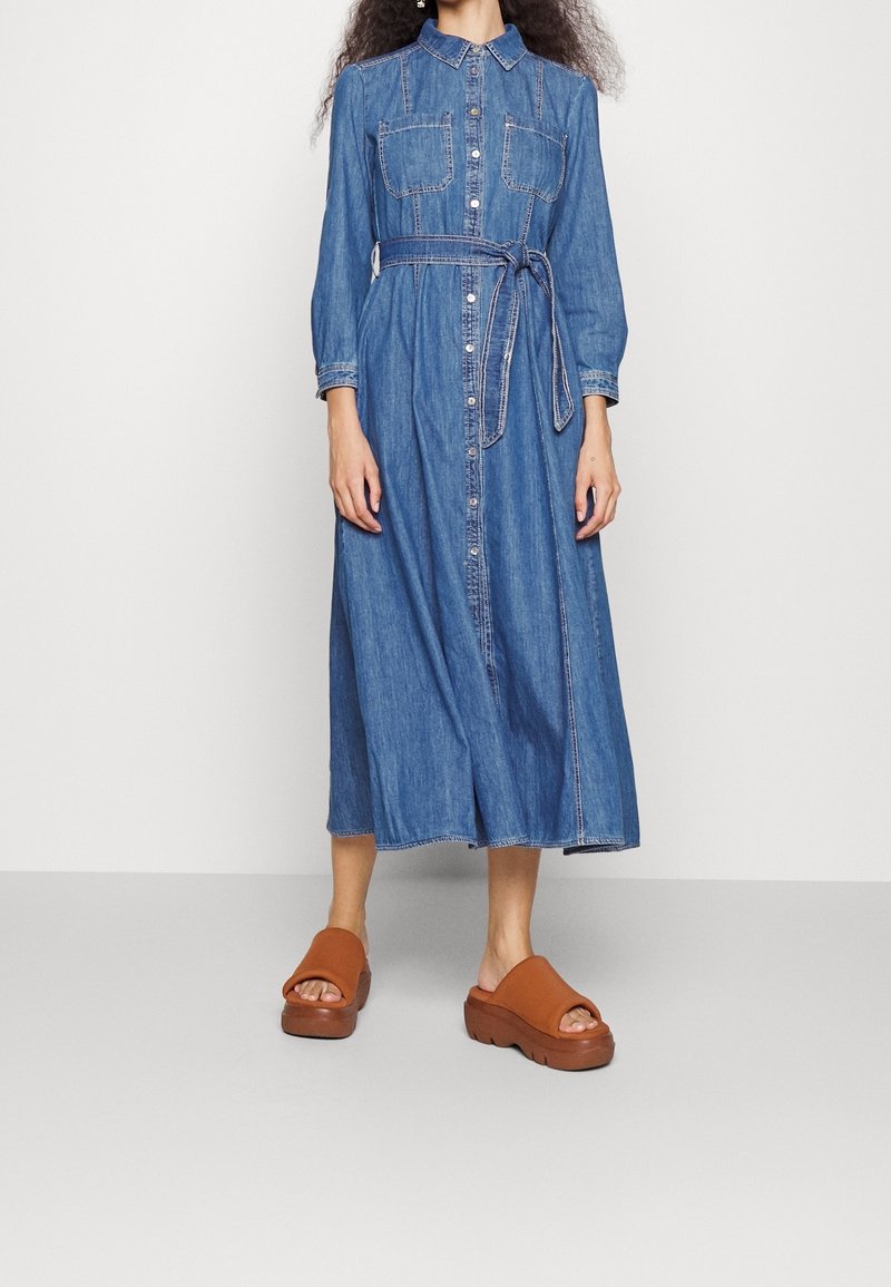 Denim shirt dress, long sleeves, button-down front, two chest pockets, waist tie, mid-calf length, finished with contrasting stitching; worn with brown platform sandals.