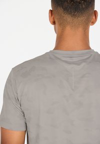 Grå atletisk t-shirt med ett strukturerat, andningsbart tyg och ett subtilt kamouflage-mönster samt en kort, rund halsringning.