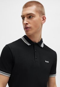 Polo noir en tissu texturé, avec un col à rayures grises et une patte de boutonnage à trois boutons. Présente un petit logo blanc "BOSS" sur la poitrine.