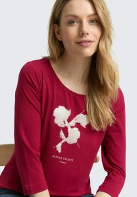Rotes Langarmshirt mit einem weißen grafischen Design von Blumen und dem Text "ALPINE ESCAPE". Weiches Material mit Rundhalsausschnitt.