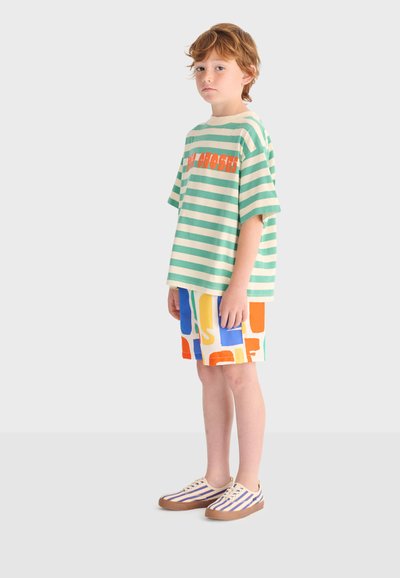 Junger Junge steht vor einem einfarbigen Hintergrund und trägt ein grün-weiß gestreiftes Shirt mit orangefarbenem Text, bunte gemusterte Shorts und gestreifte Sneakers.