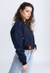 La giacca bomber navy presenta una chiusura con zip, polsini elasticizzati e una tasca frontale, realizzata in un materiale liscio con un dettaglio etichetta arancione.