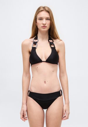 Ung kvinde med langt blondt hår iført en sort bikini med ring- og firkantede metaldetaljer på stropperne, stående mod en enkel lys baggrund.