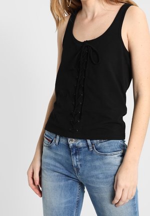 Femme portant un top noir sans manches avec un détail de laçage à l'avant et un jean délavé bleu clair, se tenant devant un fond uni.