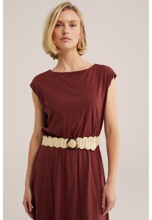 Femme blonde portant une robe bordeaux sans manches avec une large ceinture beige tissée avec une boucle ronde, debout devant un fond uni.