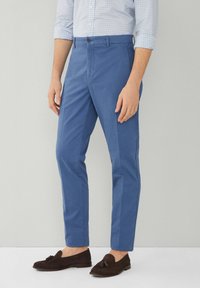 Man in strakke blauwe broek, lichtblauw geruit overhemd met opgerolde mouwen en donkerbruine tassel loafers tegen een effen achtergrond.