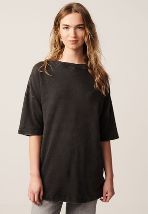 Giovane donna con capelli biondi ondulati che indossa una maglietta nera larga oversize e pantaloni grigio chiaro, in piedi davanti a uno sfondo semplice.