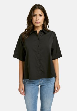 Black collared short-sleeve shirt with a button front, made of smooth fabric. Worn with light blue denim, featuring a relaxed fit.