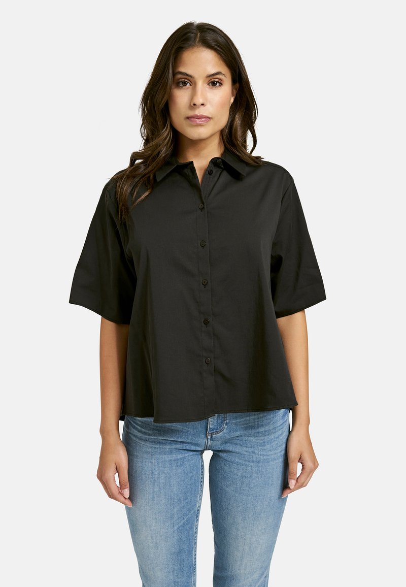 Black collared short-sleeve shirt with a button front, made of smooth fabric. Worn with light blue denim, featuring a relaxed fit.