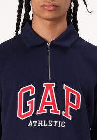 Sudadera de cremallera azul marino con un cuello alto, que presenta letras grandes rojas de "GAP" y "ATHLETIC" en blanco debajo. Textura suave.