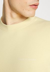 Sudadera de color amarillo claro con cuello acanalado, hecha de una tela suave. Presenta un sutil logo impreso en blanco en el pecho.