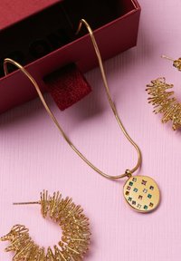 Collar de cadena de serpiente dorada con un colgante redondo que presenta piedras preciosas cuadradas de colores. Acompañado de pendientes espirales dorados sobre un fondo rosa.