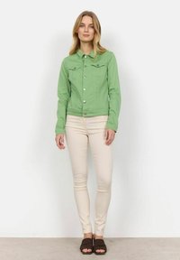 Grön denimjacka med knäppning, två bröstfickor och krage, bärs över ljusbeiga byxor och bruna slidesandaler.