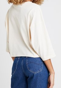 Cremefarbener, verkürzter Sweatshirt mit weiten Ärmeln, weicher Textur und rundem Ausschnitt. Kombiniert mit hoch taillierten blauen Jeans mit herzförmigen Taschen.